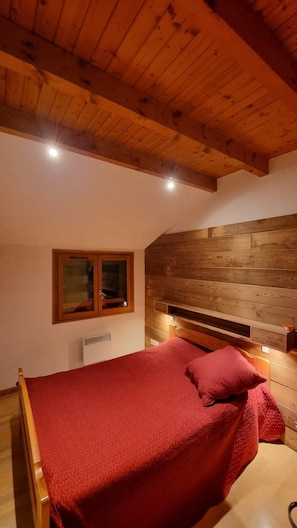 A bedroom with a wooden ceiling, a bed with a red blanket, a window, and a built-in shelf.