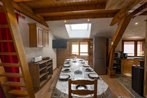A dining area with a long table set for four, wooden beams on the ceiling, a television mounted on the wall, and a kitchen area with a refrigerator and microwave.