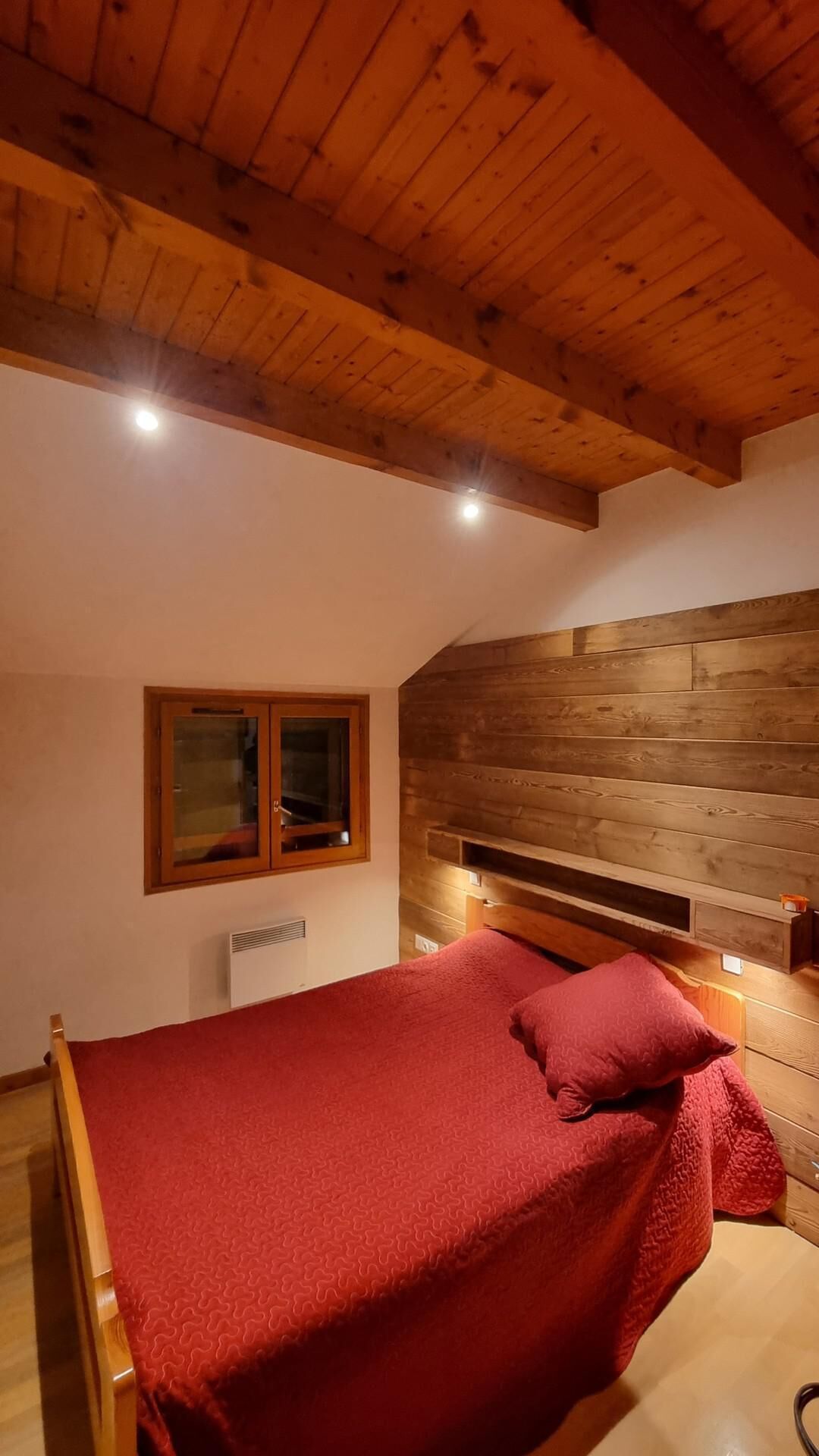 Een slaapkamer met een houten plafond, een bed met een rode deken, een houten hoofdbord en een raam met houten kader.