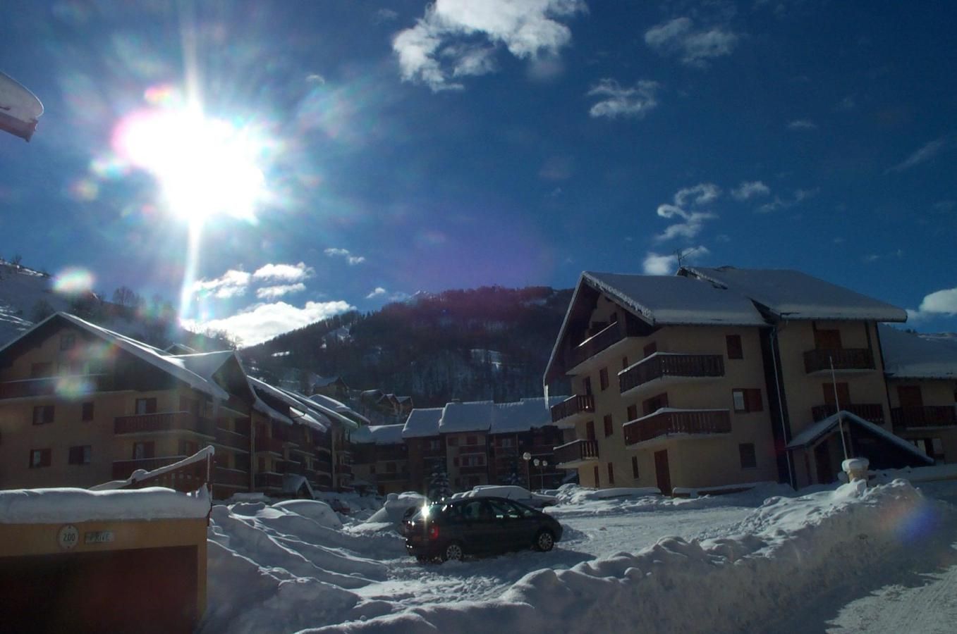 A snow-covered building with multiple balconies, a parked car, and a mountain in the background.