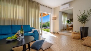 A modern living room with a blue sofa, a round table with a bottle of wine, and a bowl of fruit. There is a large potted plant and a view of the outdoors through a glass door. - Luxury Villa Essencia Milna Brac with Heated Pool (Milna (Brac))