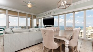 A spacious living area with a large window, a ceiling fan, a dining table with chairs, and a flat-screen TV.