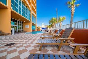 A poolside area with lounge chairs and a checkered floor, overlooking a building with large windows and palm trees. - Phoenix Gulf Shores 2001 4 Bedroom Condo (Gulf Shores)