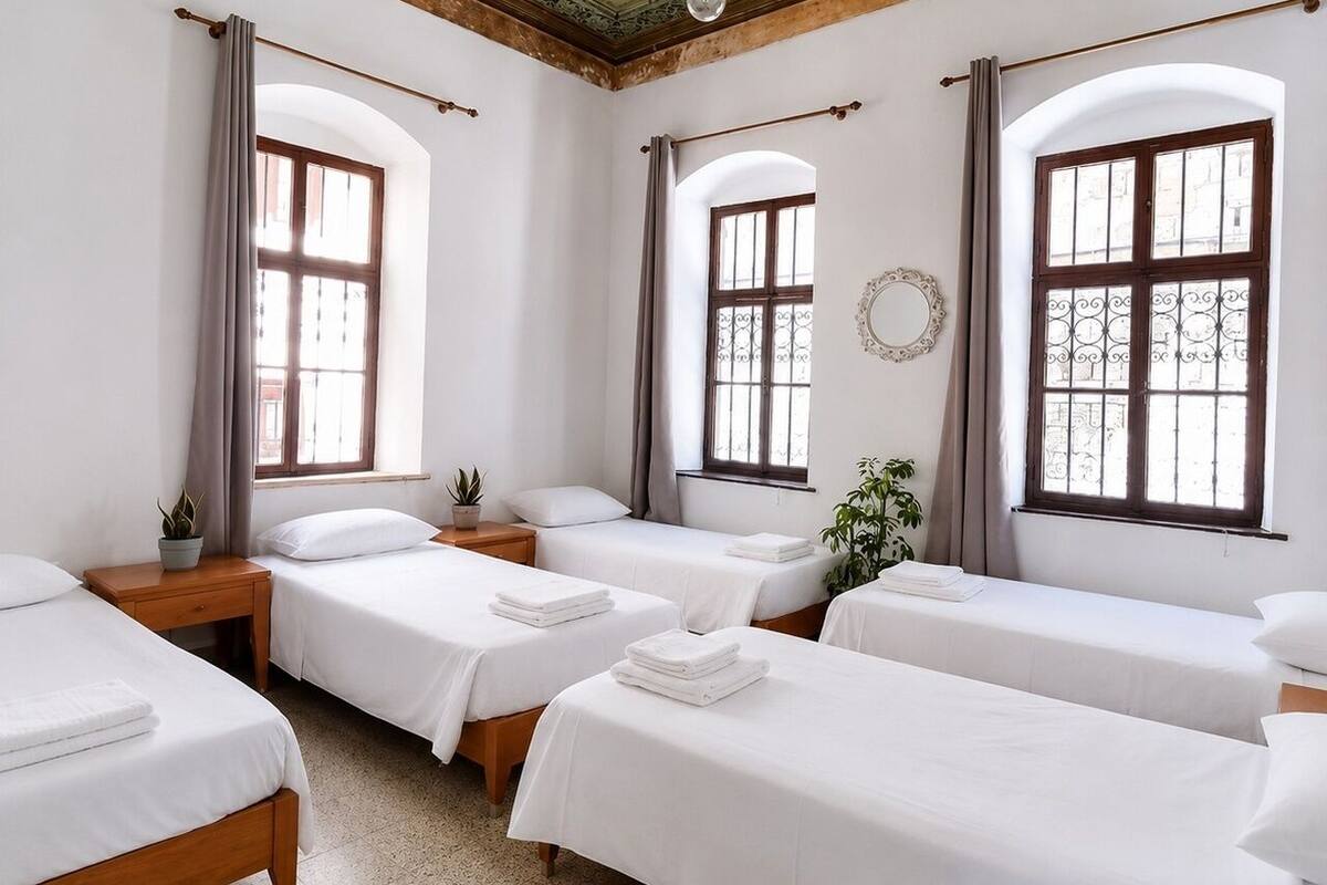 A room with four single beds, wooden headboards, a round mirror, and two windows with curtains.
