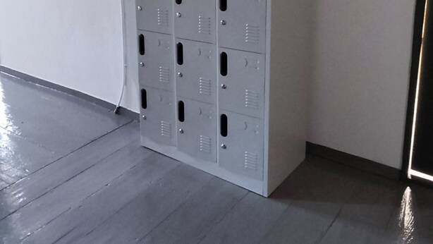 Een rij grijze metalen locker in een hoek van een ruimte met een grijze vloer en witte muren.
