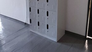 A row of grey metal lockers in a corner of a room with a grey floor and white walls. - Rhodes Youth Hostel (Rhodes)