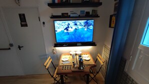 Un comedor con una mesa dispuesta para dos personas, un televisor montado en la pared y una estantería sobre la mesa con libros y objetos decorativos.