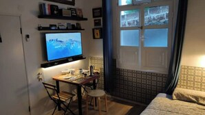 Una habitación pequeña con una televisión en un estante, un escritorio con una silla, una cama y una ventana con cortinas.
