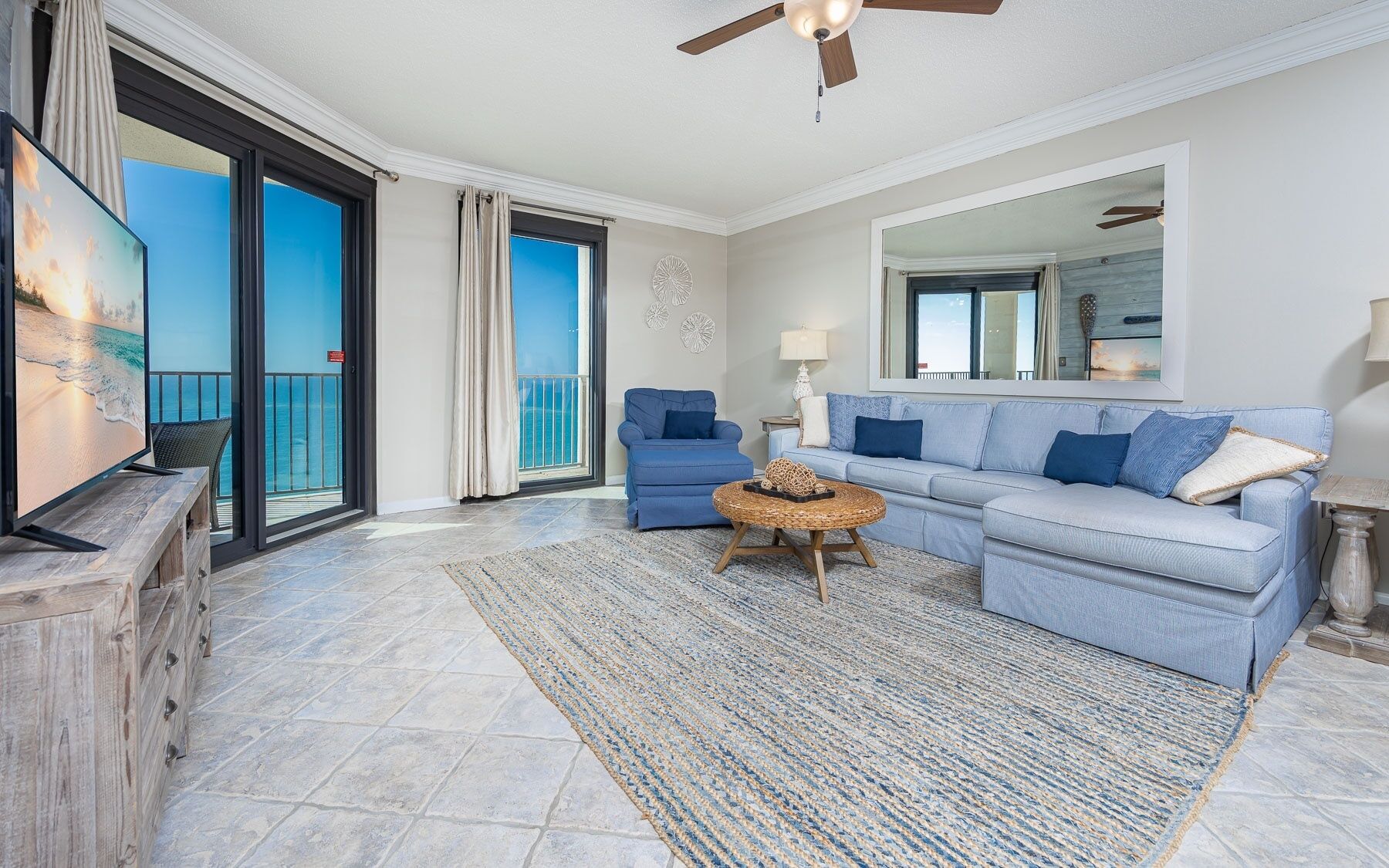 A living room with a sofa, a coffee table, and a television. There is a balcony with a view of the ocean.