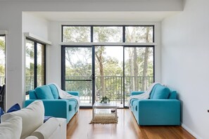 A modern living room with two teal sofas, a white sofa, and a coffee table. Large windows provide a view of trees and a balcony. - Family Retreat, Bay Views, 5 Mins Bagnall's Beach (Nelson Bay)