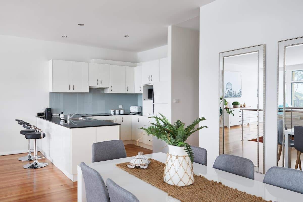 A modern kitchen with white cabinets, a black countertop, and a central island. There are two bar stools and a dining table with chairs. A potted plant is on the island.