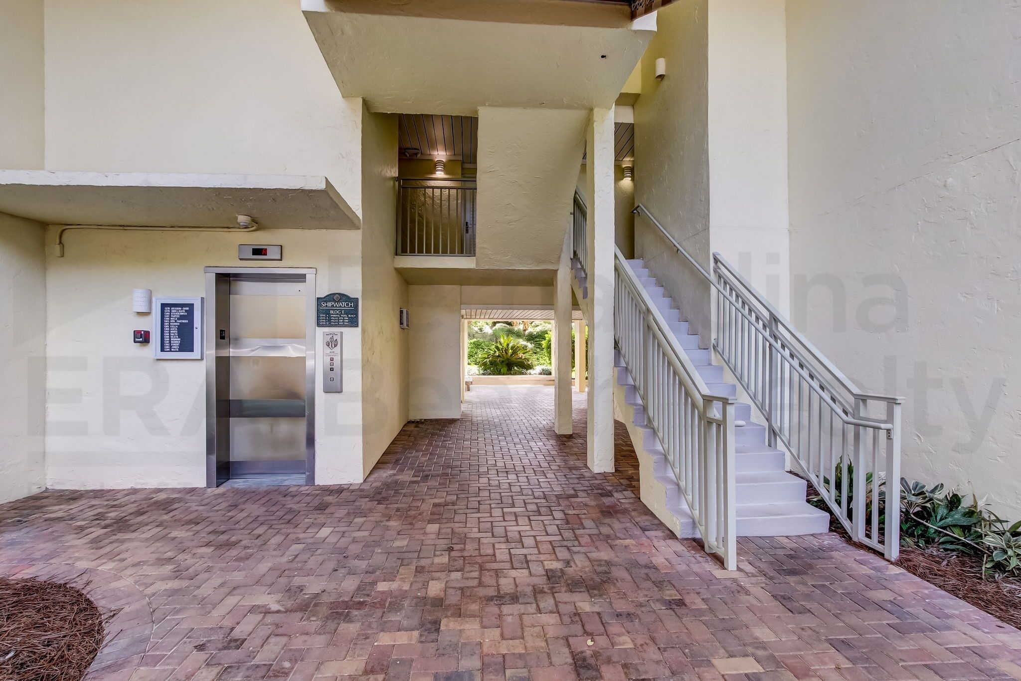 A covered walkway with a brick floor, a metal staircase, and a door with a sign.