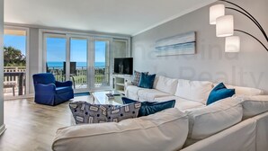 Un salon moderne avec un canapé blanc, un fauteuil bleu et un pouf à motifs. Un téléviseur à écran plat est fixé au mur, et il y a un balcon avec vue sur la mer.