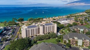 An aerial view of a coastal area with a large hotel featuring a parking lot, solar panels, and a view of the ocean and mountains.