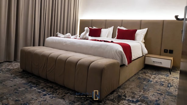 A hotel room with a large bed, a bedside table, a footstool, and a carpeted floor.
