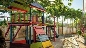 A colorful playground with a slide and climbing structures, surrounded by palm trees and a brick wall. - Chez soi Loin de Chez soi (Anse Royale)