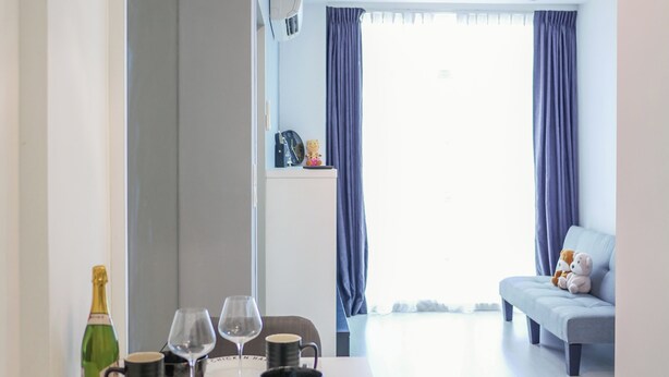 A modern dining area with a table set for two, a decorative pendant light, and a view of a living room with a sofa and curtains.
