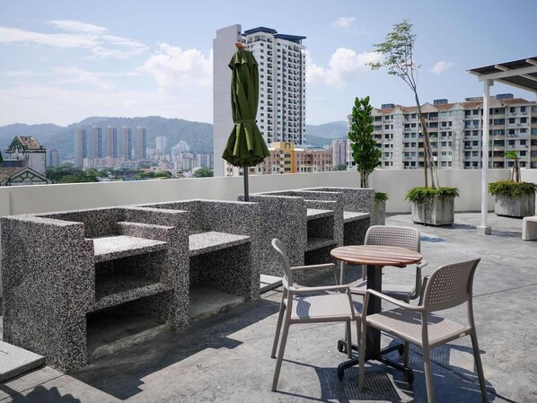 A rooftop terrace with outdoor seating, a green umbrella, and a view of a cityscape with buildings and mountains in the background.