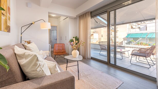 Ein modernes Wohnzimmer mit einer Couch, einem kleinen Tisch und einer Stehlampe. Ein großes Schiebeglasfenster führt auf einen Außenbalkon mit Stühlen und einem Tisch.