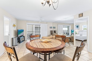 A spacious living area with a round wooden table, comfortable seating, a television, and a balcony with a view. - Crescent Arms by Beachside Management (Siesta Key)