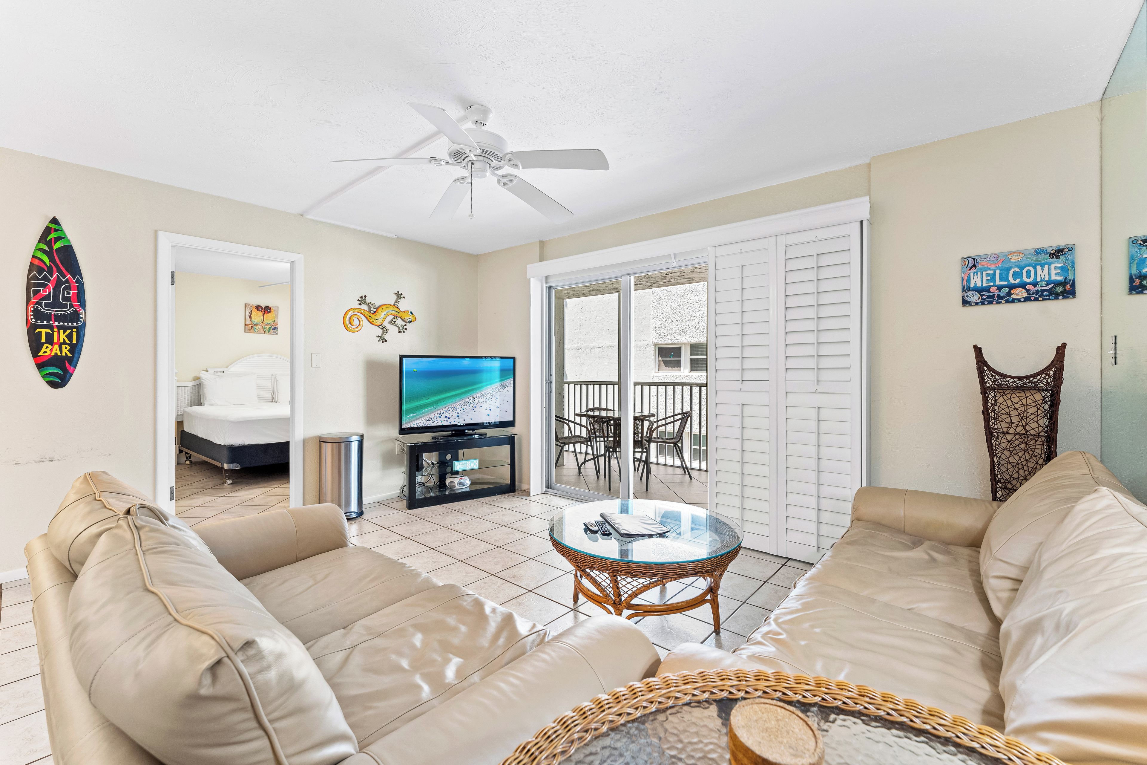 A living room with beige sofas, a glass-top coffee table, a ceiling fan, a TV, and a balcony with a view.