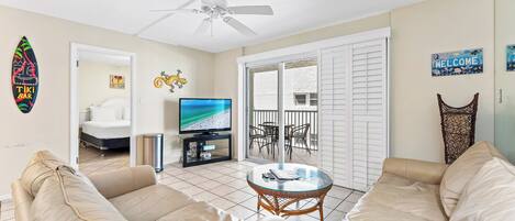 A living room with beige sofas, a glass-top coffee table, a ceiling fan, a TV, and a balcony with a view.
