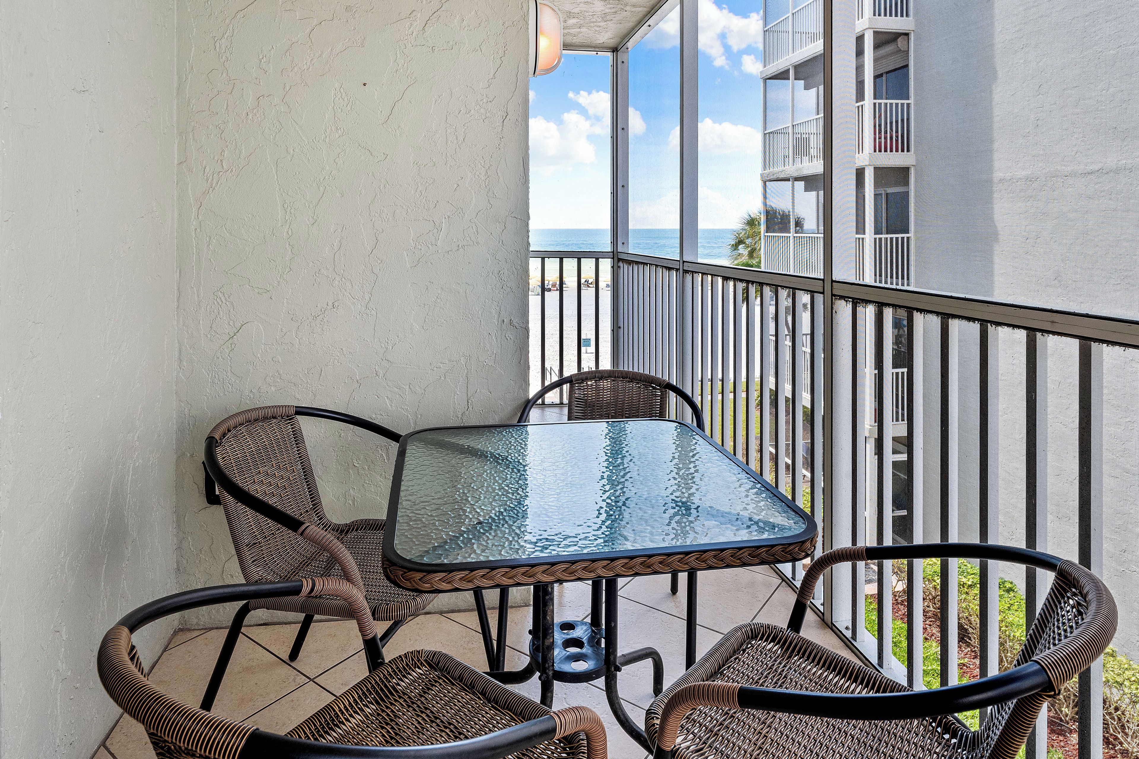 A balcony with wicker chairs and a glass table, overlooking a beach and a building with balconies.