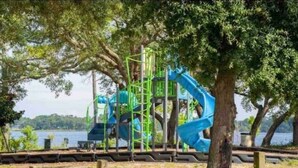 A playground with a blue slide and climbing structure, surrounded by trees and a body of water. - Cute cottage near the Navy base (Pensacola)