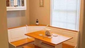 A small, cozy room with a wooden bench seating area around a central table, a window with blinds, and a cabinet with glass doors. - Cute cottage near the Navy base (Pensacola)