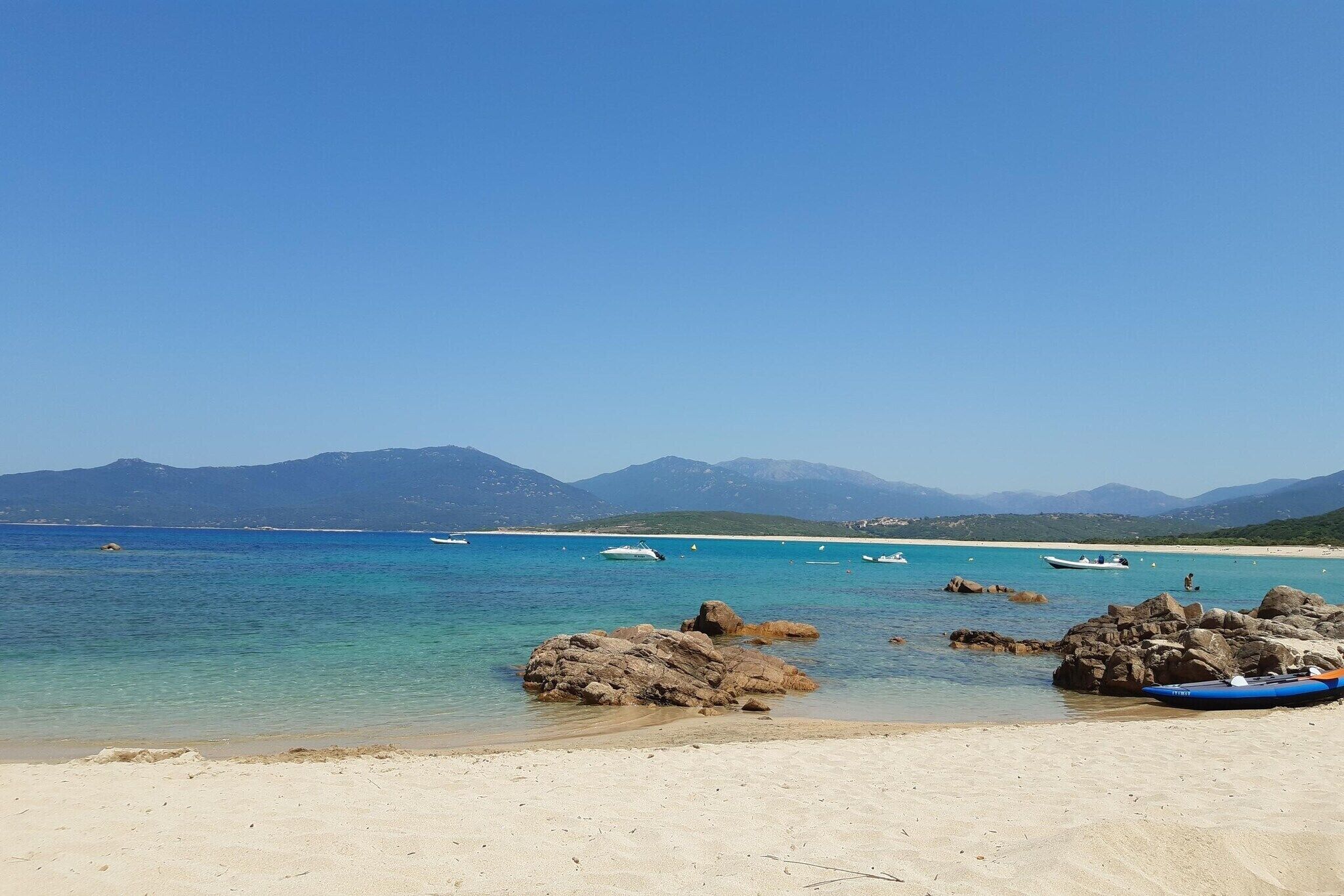 A beach with clear water, rocks, and boats. Mountains are visible in the background.