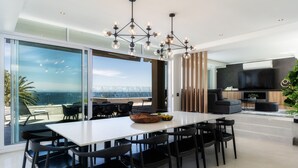 A modern dining area with a large table and chairs, a chandelier, and a view of the ocean. - 26OnFirst: UPS, Sea Views & Personalised Service (Cape Town)