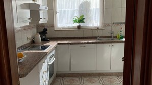 Cozinha com armários brancos, janela com planta e chão ladrilhado.
