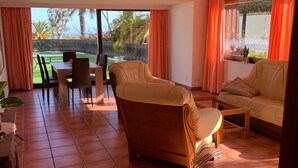 Uma sala de estar ampla com sofá, mesa de jantar e cadeiras. Há vista para um jardim através de grandes janelas.