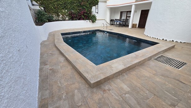 Piscina retangular com borda de pedra, cercada por um pátio com chão ladrilhado e uma parede branca.
