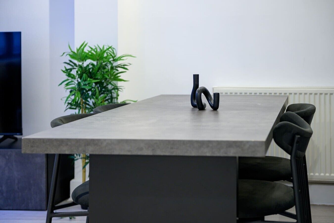 A modern conference room with a rectangular table, black chairs, a potted plant, and a radiator.