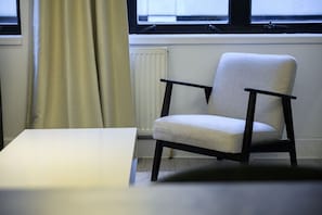 A chair with a light-colored cushion and dark wooden armrests, positioned near a window with a view of the outside.