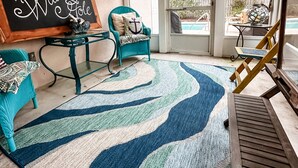 Un espace patio accueillant avec une chaise de couleur turquoise, une table en bois et un tapis bleu et blanc. Un panneau noir est accroché au mur et, de là, on aperçoit une piscine à l’extérieur.