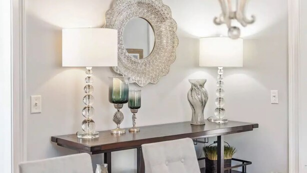 Un coin repas avec une table en bois foncé, deux chaises blanches, un miroir décoratif et deux lampes murales dotées d’abat-jour en verre transparent.