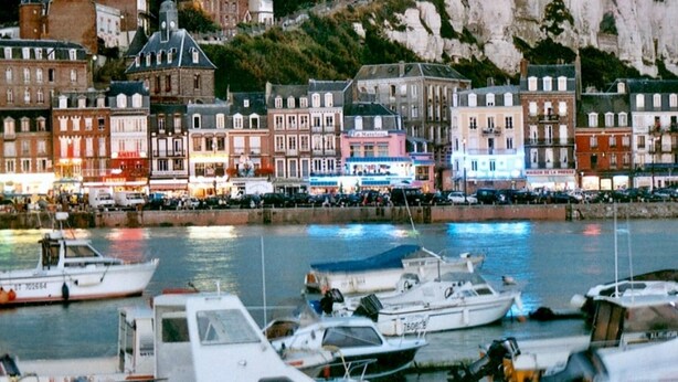 A marina with boats docked, a waterfront promenade with buildings, and a cliff in the background.