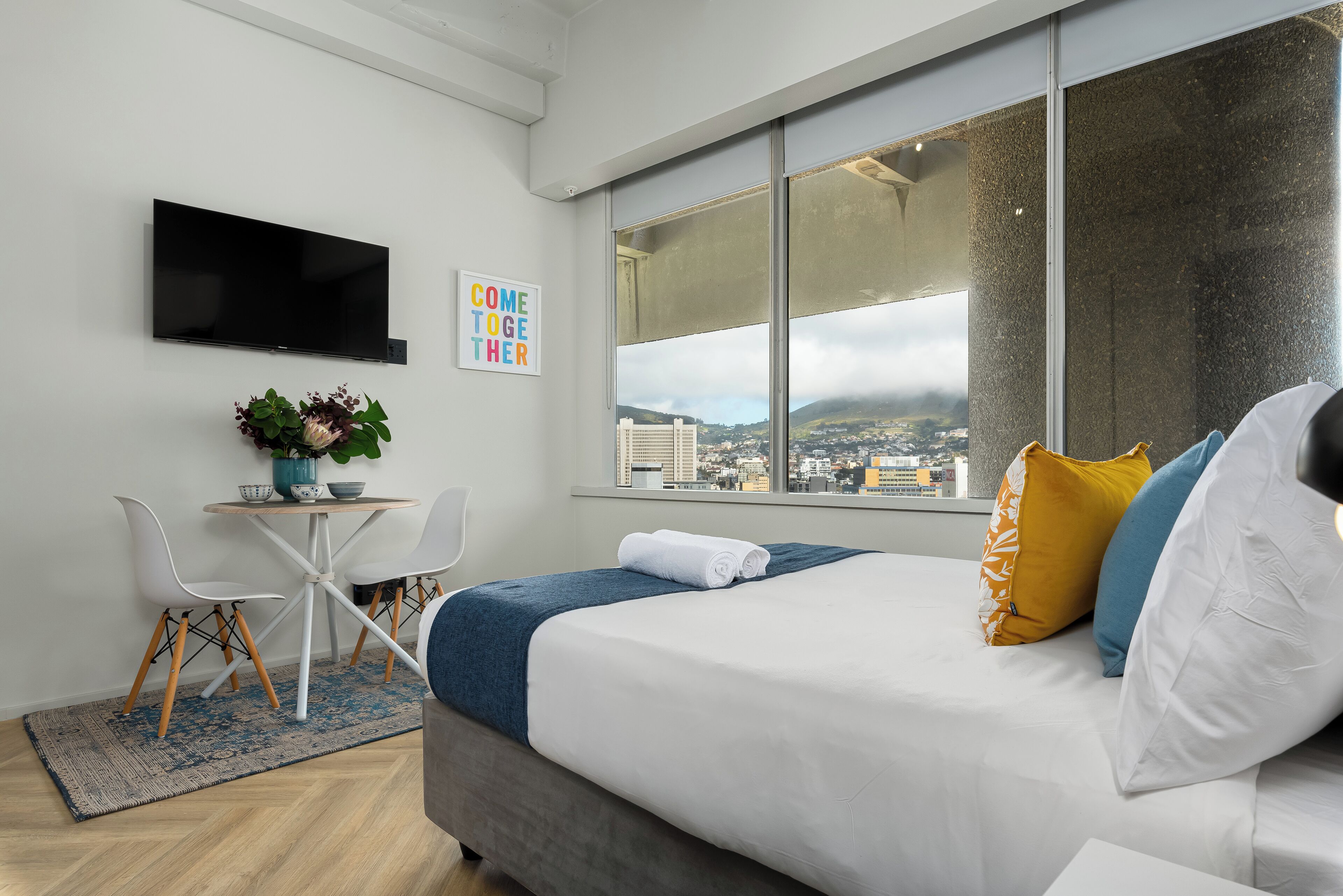 Ein modernes Hotelzimmer mit einem Bett, einem Schreibtisch mit Stühlen, einem Fernseher und einem großen Fenster mit Blick auf die Stadt.