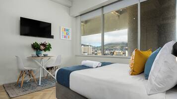 Ein modernes Hotelzimmer mit einem Bett, einem Schreibtisch mit Stühlen, einem Fernseher und einem großen Fenster mit Blick auf die Stadt.