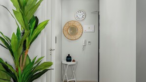 Un pasillo con una planta en maceta, un reloj de pared, un espejo decorativo, una mesita con un jarrón y un termostato fijado a la pared.