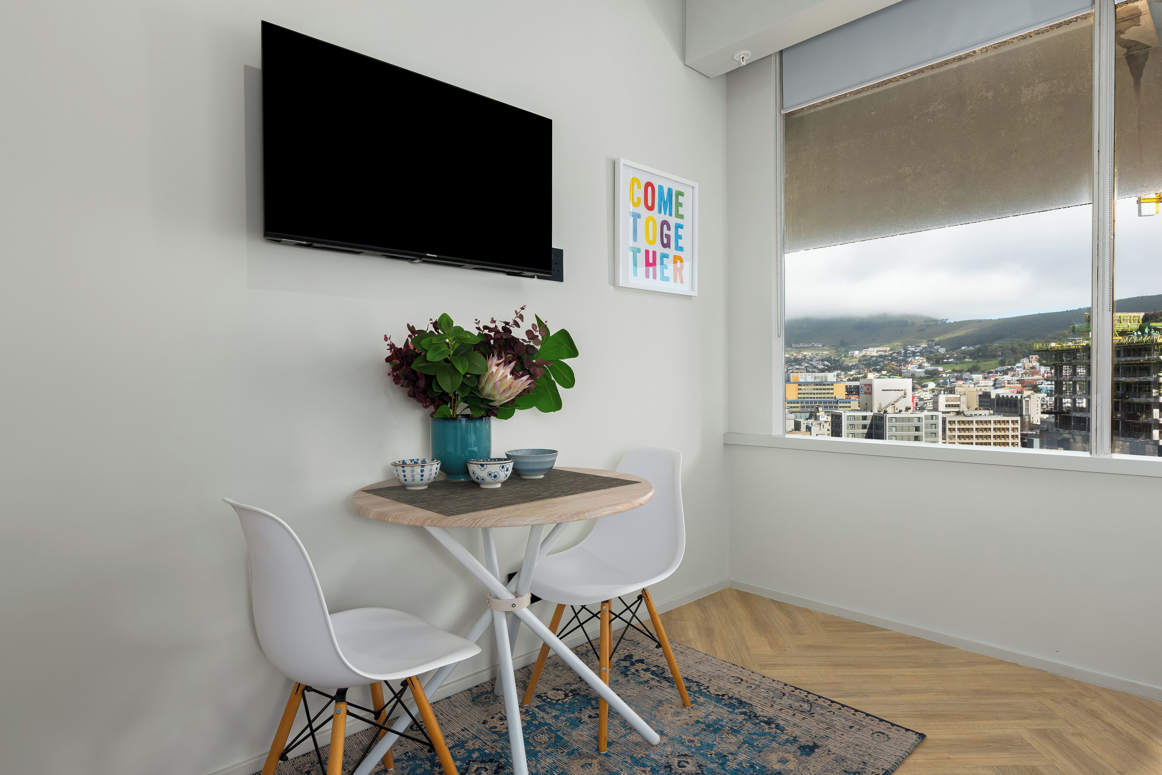 A modern room with a TV mounted on the wall, a small table with chairs, a city view through a large window, and a "COME TOGETHER" sign.