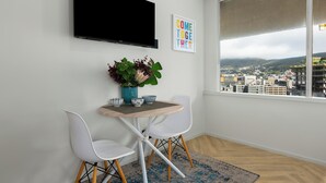 Una habitación moderna con televisor montado en la pared, una pequeña mesa con sillas, vista a la ciudad a través de un ventanal grande y un cartel que dice 'COME TOGETHER'.