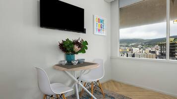 A modern room with a TV mounted on the wall, a small table with chairs, a city view through a large window, and a "COME TOGETHER" sign.