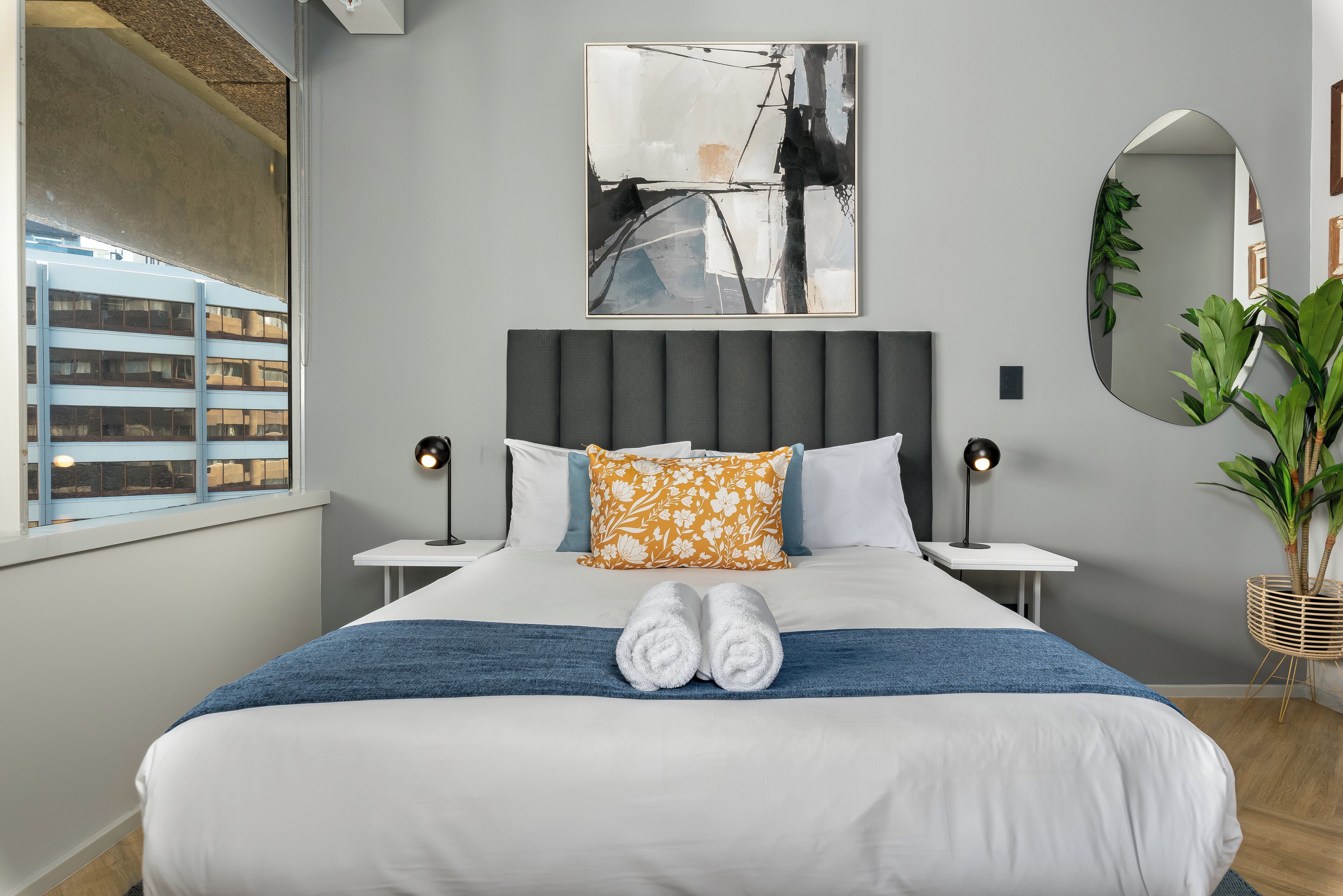 Ein ordentlich bezogenes Bett mit weißer und blauer Bettwäsche, einem Blumenkissen und zwei aufgerollten Handtüchern.