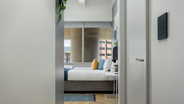 Un dormitorio moderno con cama, mesita de noche y vistas a la ciudad a través de amplias ventanas.