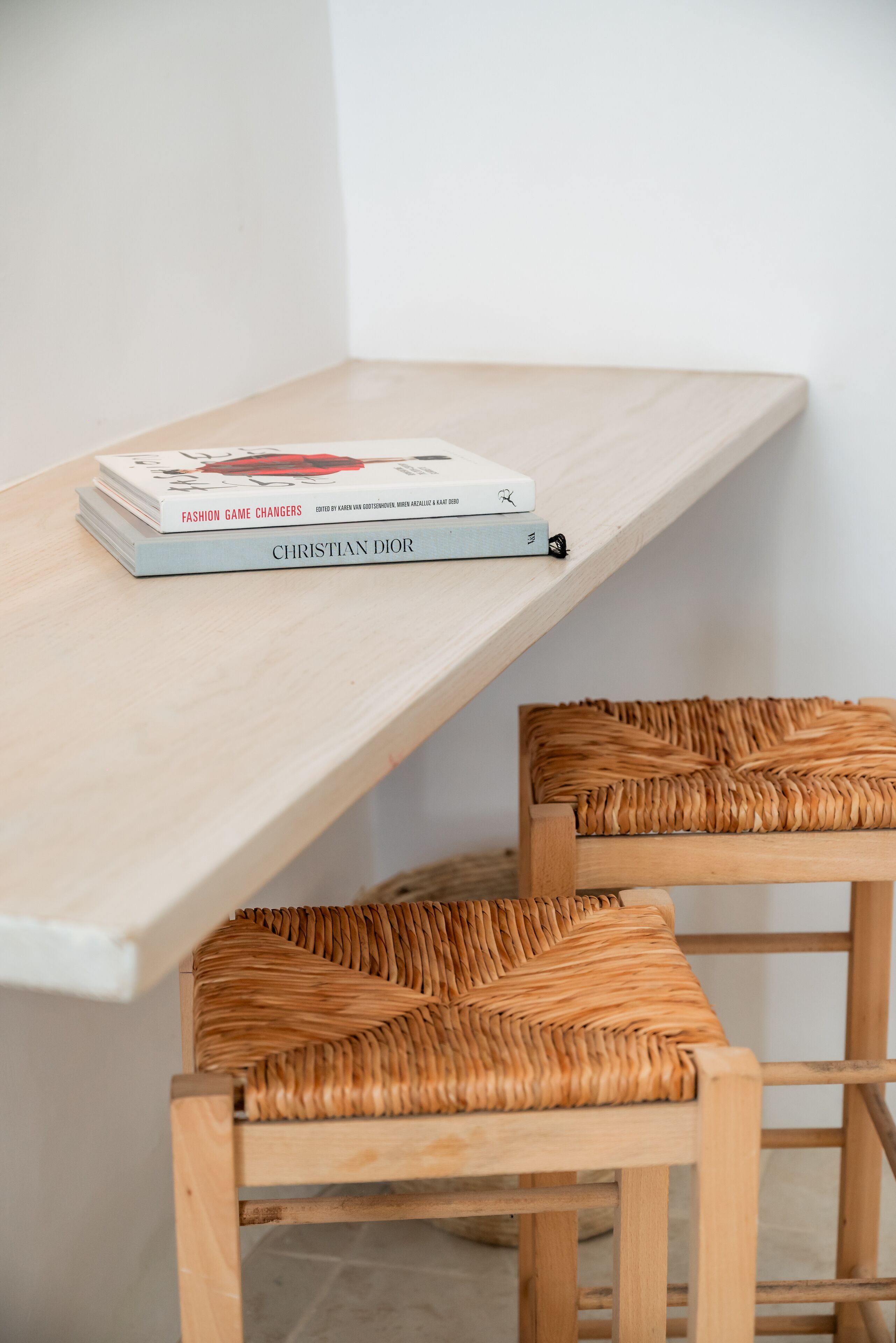 A wooden table with a stack of books titled "Fashion Game Changers" and "Christian Dior." Two woven chairs are positioned below the table.