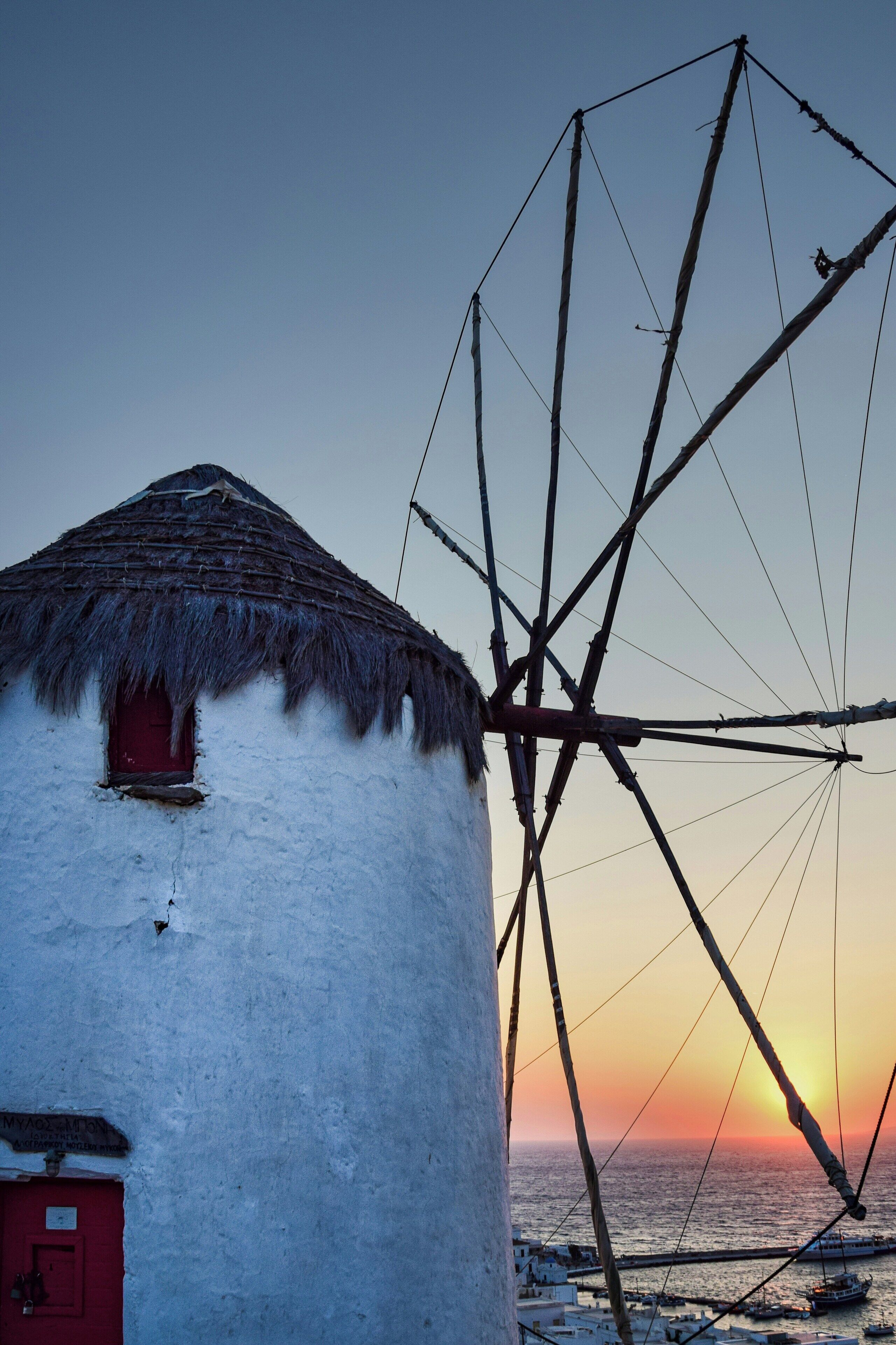 Ein traditionelles Gebäude mit einem strohgedeckten Dach, einer roten Tür, einer großen Netzkonstruktion und einem Sonnenuntergang über dem Meer.