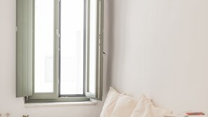 Un dormitorio minimalista con una cama, una ventana con contraventanas verdes, una planta pequeña y una mesita de noche con un libro.